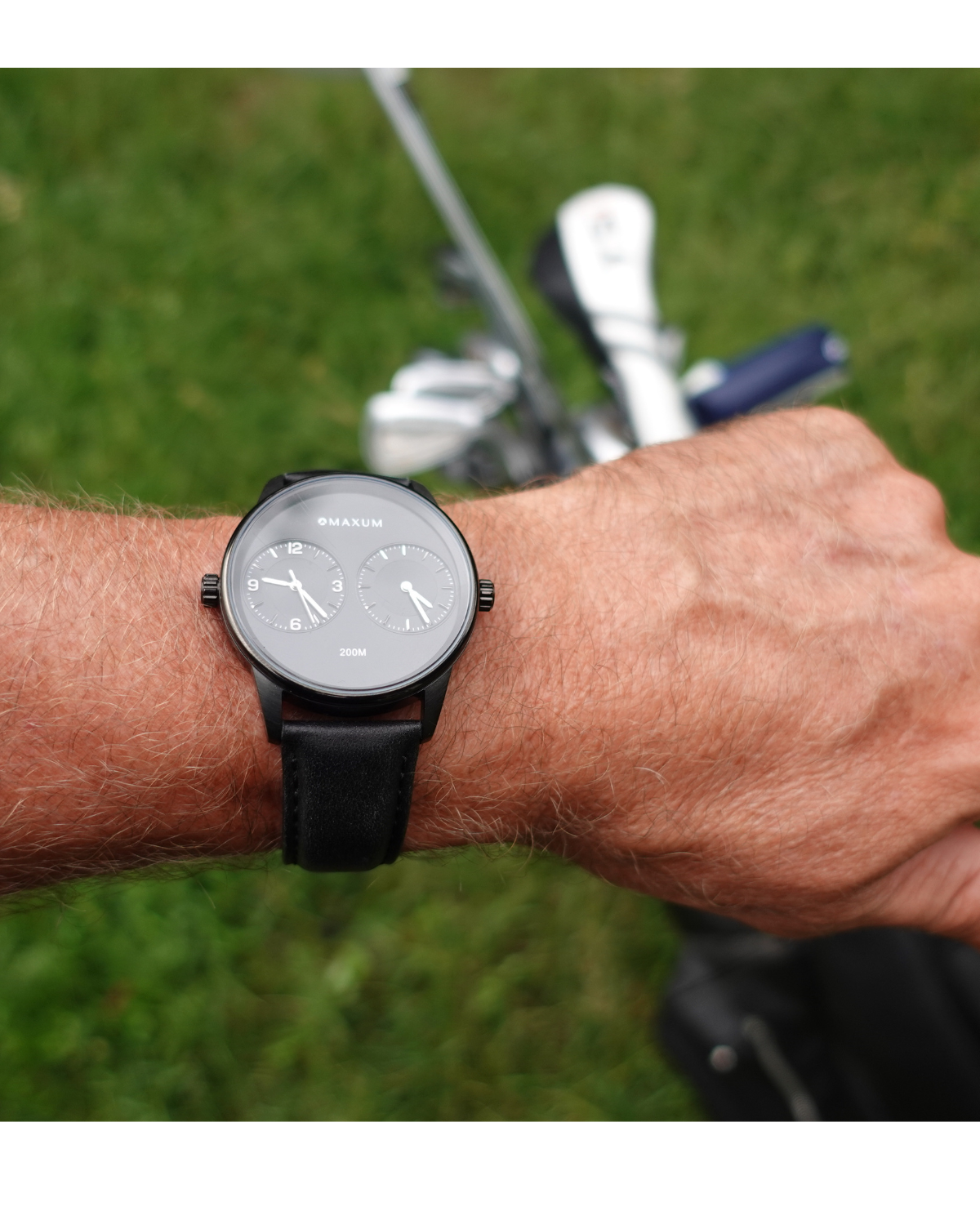 Maxum x Taylor Steele collab- black analog watch with a black dial, black case, and black leather band, branded with 'MAXUM' on the top and indicating '200M' water resistance. Person wearing on a golf course