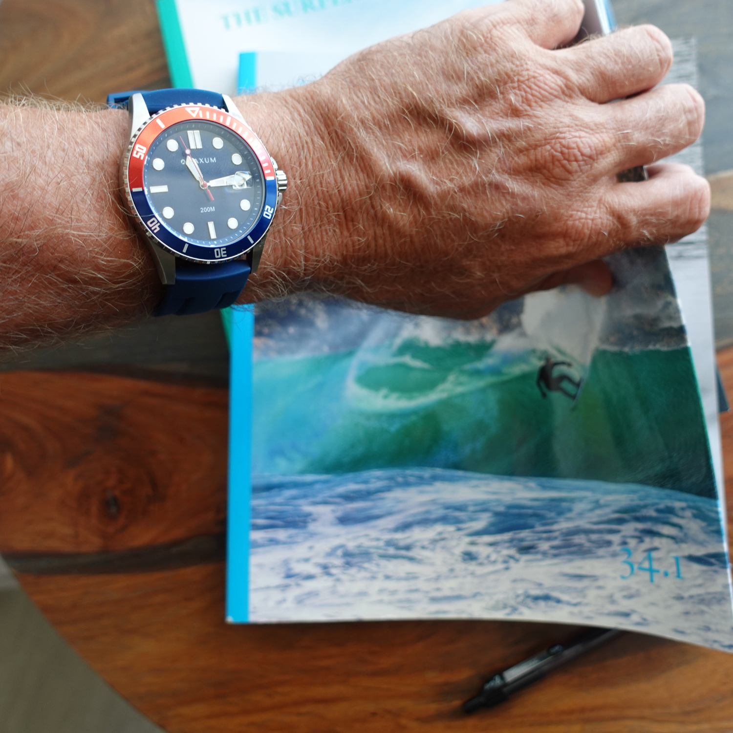 Hand wearing Maxum Prince silver-tone casing watch with a blue dial and blue silicone strap, featuring a red and blue bezel and a date display.Holding a book with a beach scene on a wooden surface.