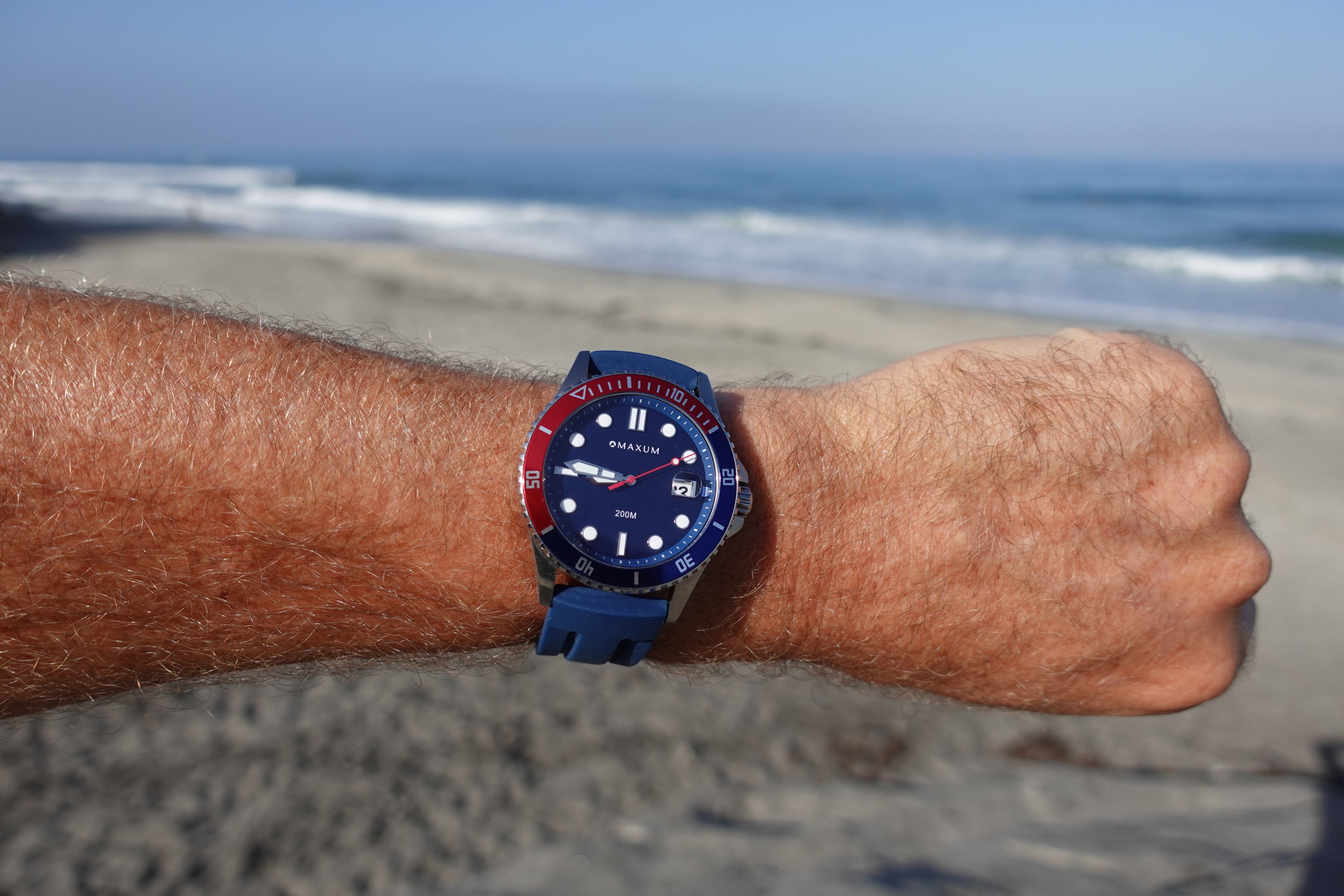 Maxum Prince silver-tone casing watch with a blue dial and blue silicone strap, featuring a red and blue bezel and a date display, on a wrist with a beach background