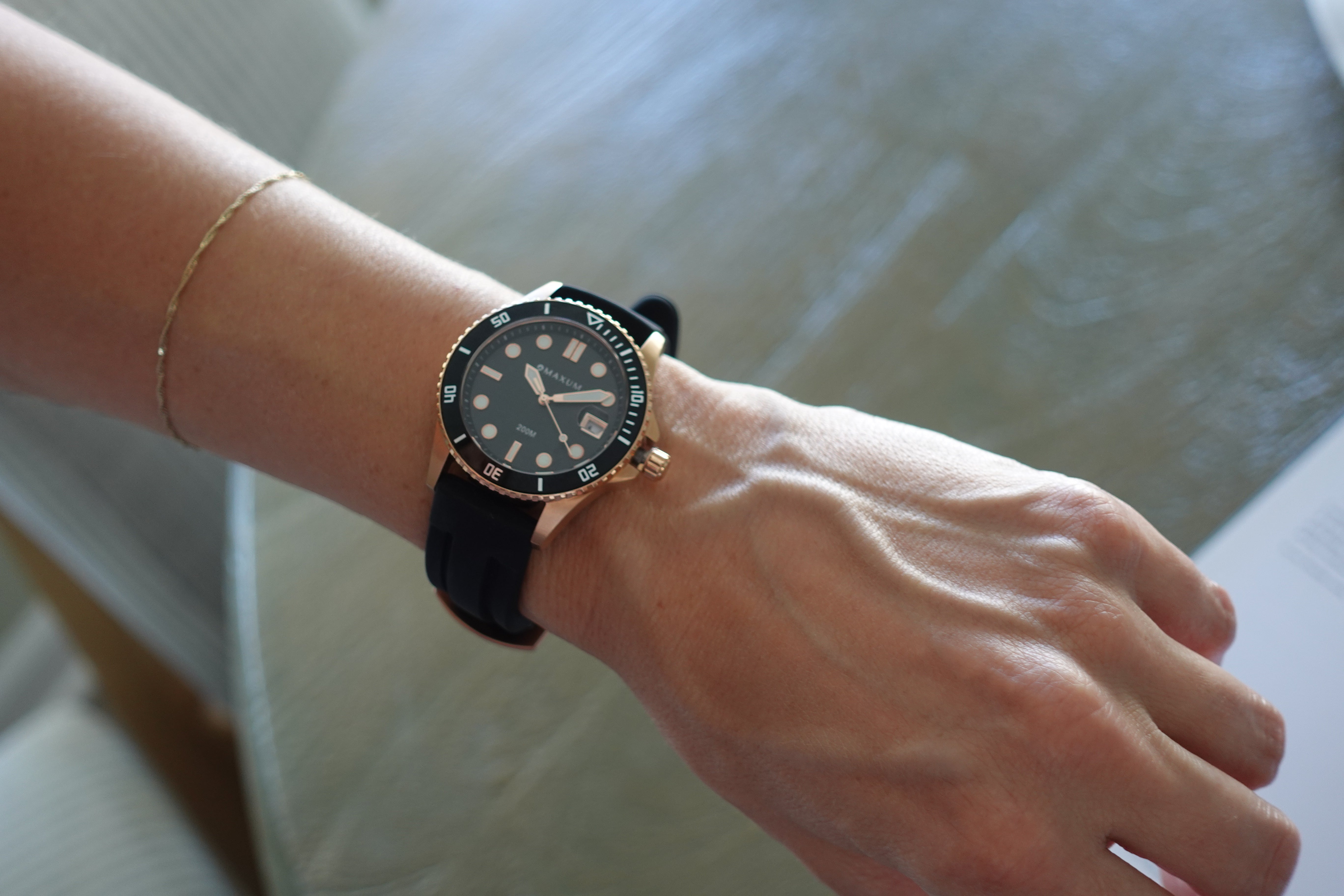 Hand wearing a Maxum Liberty watch with a black strap and rose gold case on a neutral background