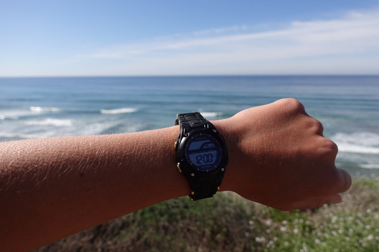 Person wearing watch with a resin strap and buckle closure. The watch has a digital display with gold-coloured accents on the casing with ocean view in the background