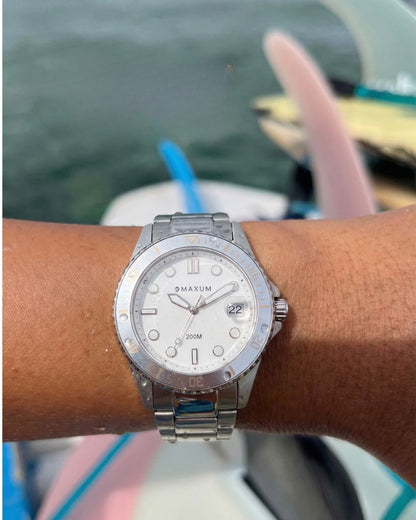 Maxum Liberty white dial watch with a silver-tone case and bracelet, featuring analog display and date function on a wrist with surfboards in blurred background