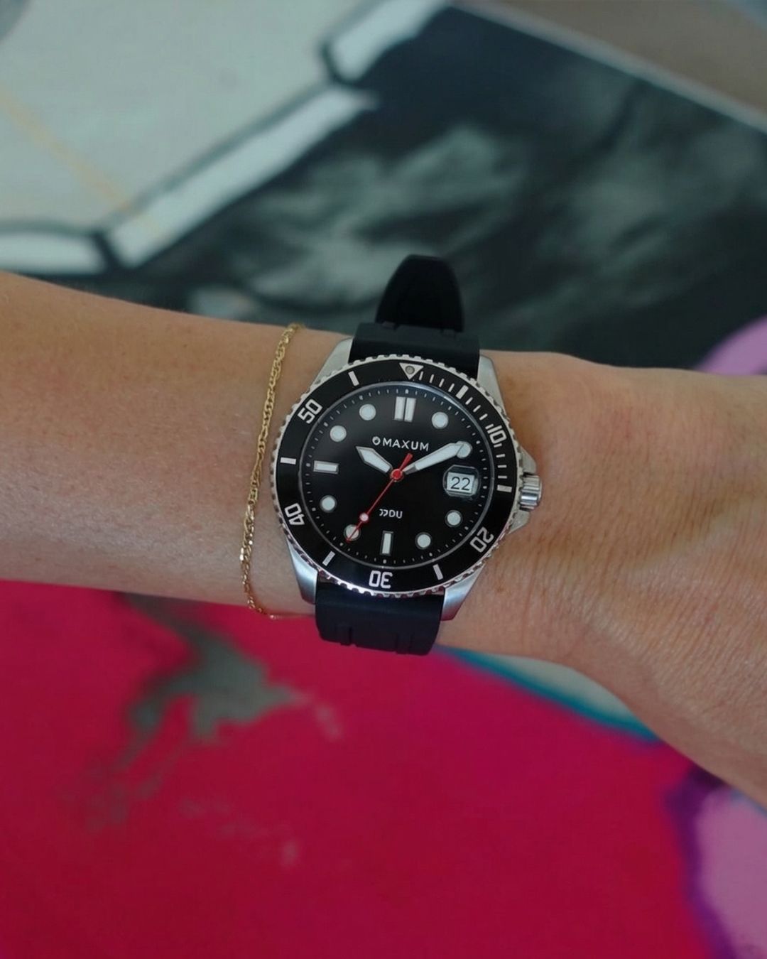 Maxum Liberty silver-tone casing watch with a black dial and black silicone strap, showing time and date.with a person's arm wearing it, over a pink surfboard blurred background
