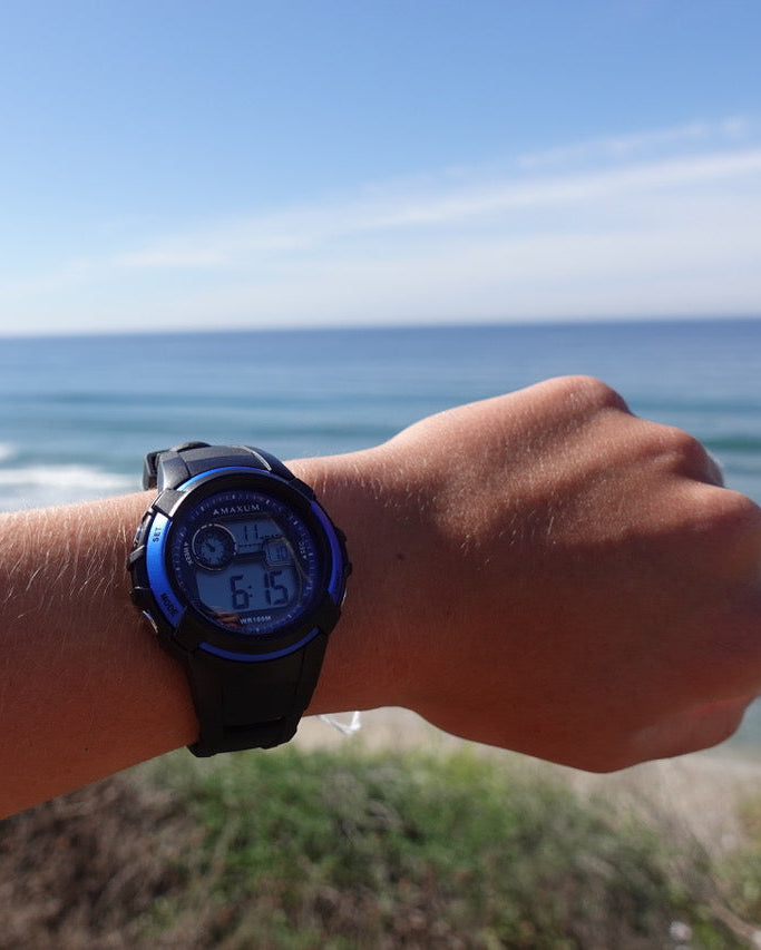 Maxum Button digital watch with a black resin strap and black case, featuring blue accents and a digital display on wrist at beach