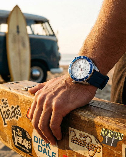Person wearing Hockey Dad x Maxum collab Base Camp watch on a wooden crate with a vintage van and surfboard in the background