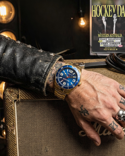 Hand wearing Hockey Dad x Maxum collab  gold watch with blue face on a textured surface, with a background featuring a gig poster 'Hockey Dad'.