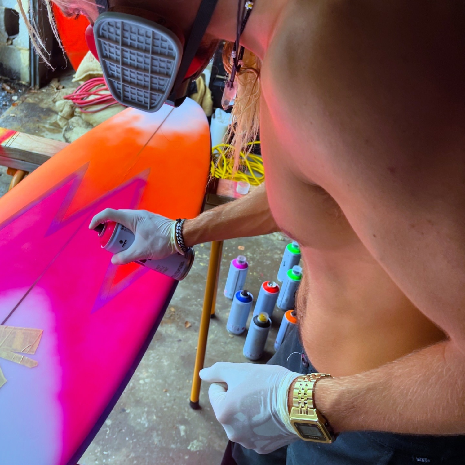Kyuss King wearing Raglan Gold Tide Watch with a black dial and a stainless steel gold-tone bracelet. The watch has a square case with a digital display showing the time and tide features. Kyuss King is  spray painting a colourful surfboard with various spray paint cans in the background.