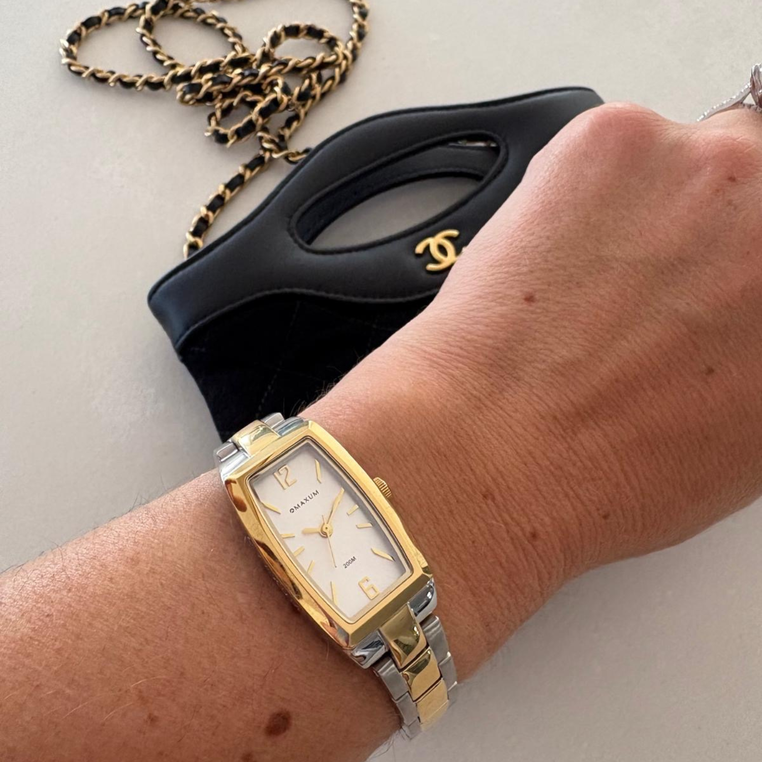 Maxum Fanta Re-issue rectangular analogue watch with a silver-tone case and a white dial, accompanied by a two-tone stainless steel bracelet with gold and silver tones, on a wrist with a black handbag featuring a gold chain in the background