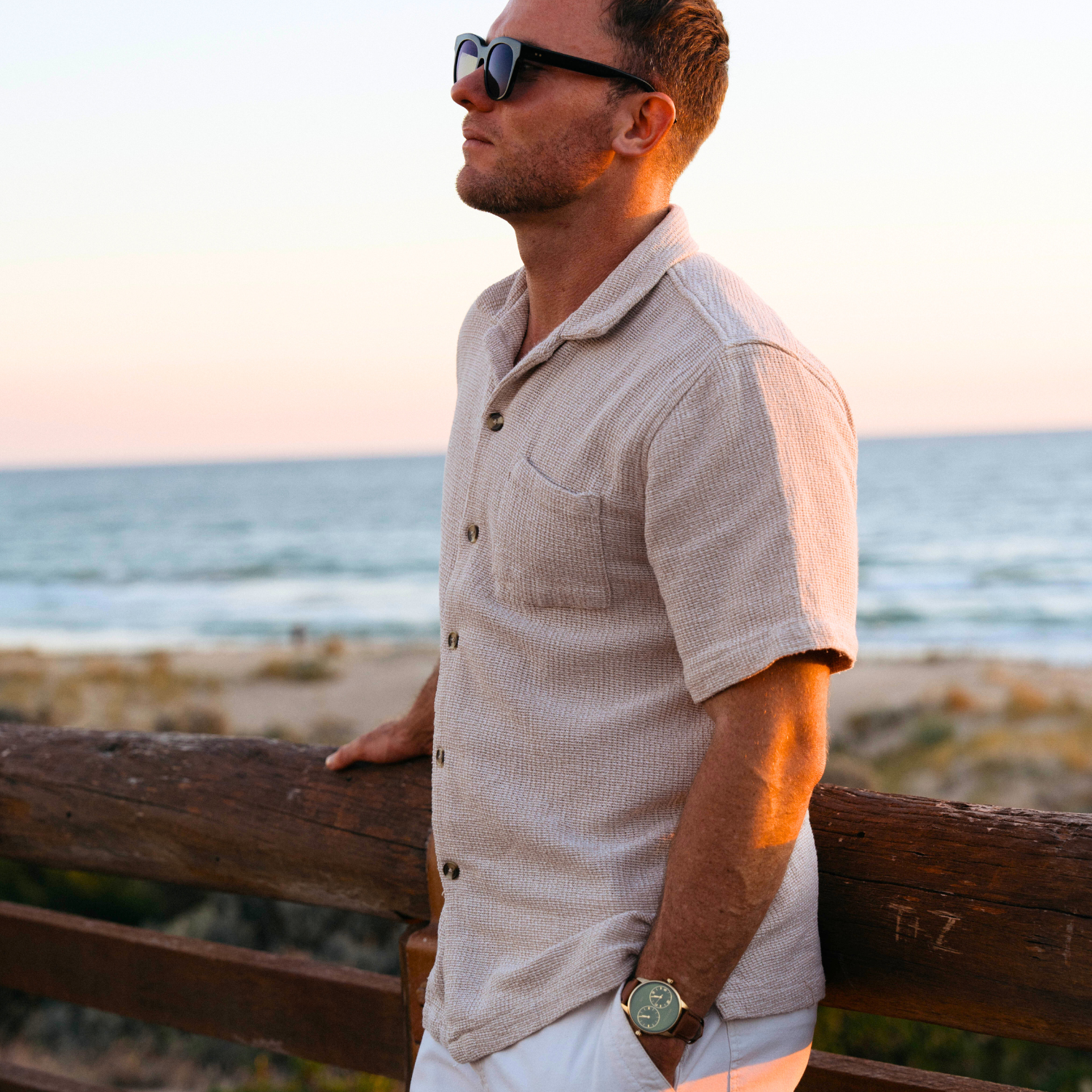 Maxum Taylor Steele Collaboration Watch with a green dial, gold-tone case, and brown leather band.Man wearing sunglasses and a beige shirt standing by a wooden railing with ocean in the background