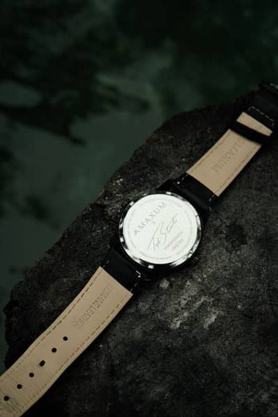 Maxum x Taylor Steele collab- black analog watch with a black dial, black case, and black leather band, back view on black rock surrounded by green water