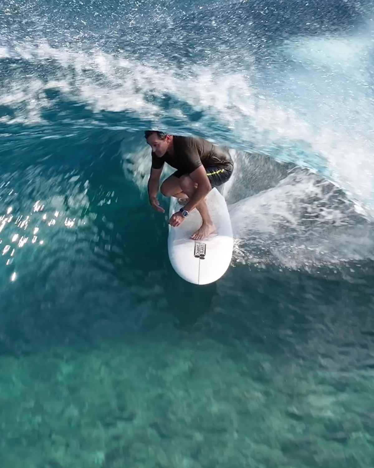 A surfer rides a curling turquoise ocean wave on a white surfboard, captured from above with bright sunlight and splashing water.