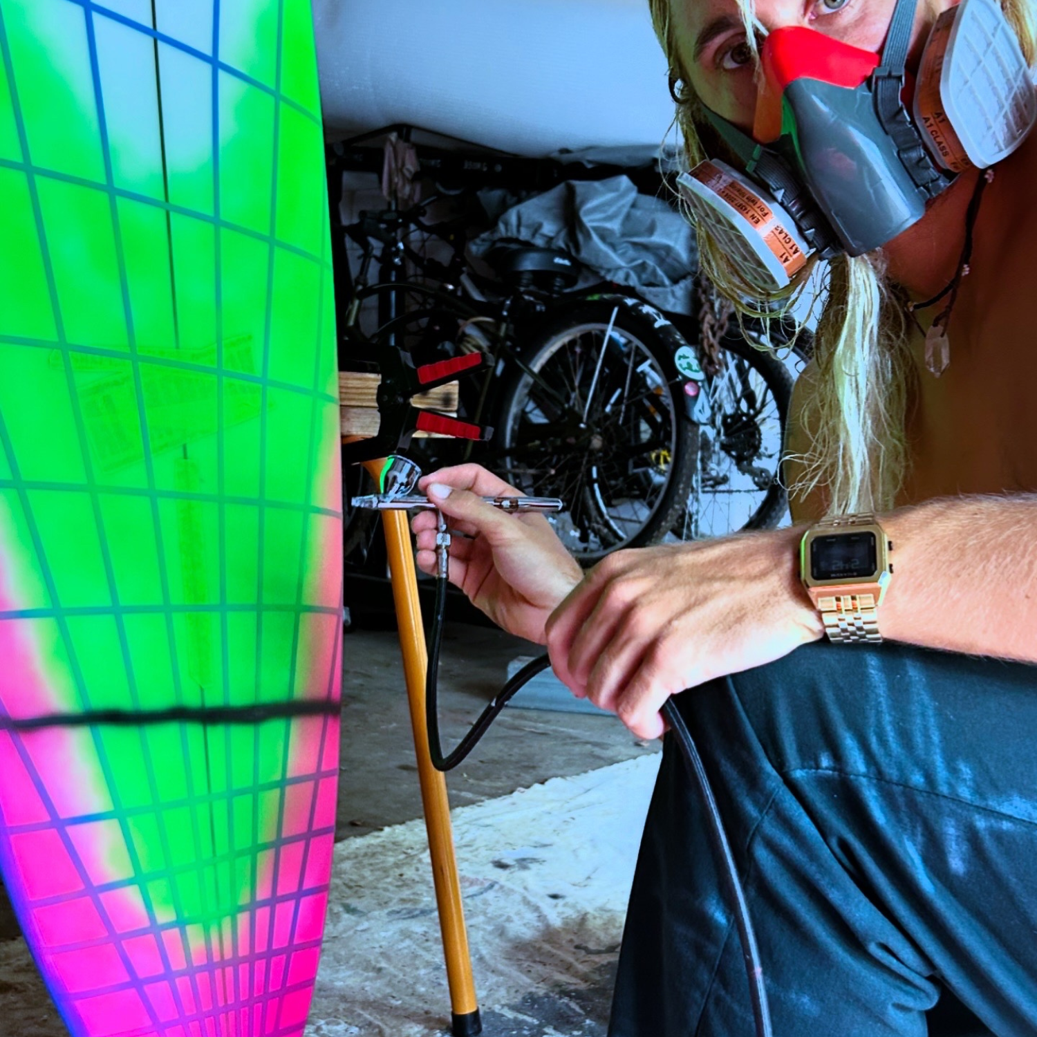Kyuss King wearing Raglan Gold Tide Watch working on a colourful surfboard in a garage setting